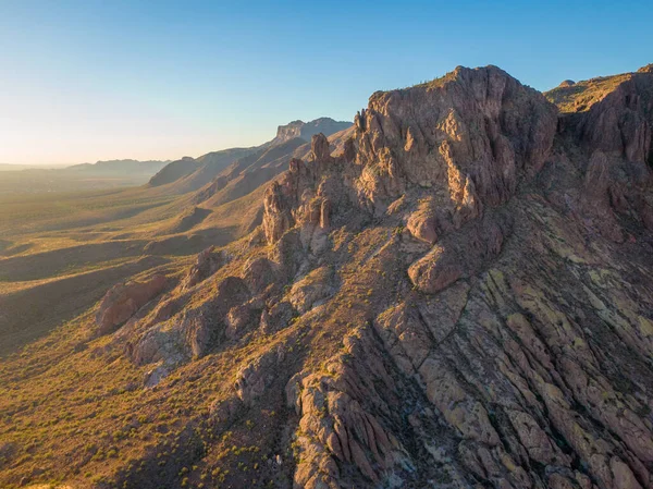 Arizona 'daki Altın Kanyon' daki engebeli Batıl inanç Dağları 'nın insansız hava aracı görüntüsü. Sıcak sabah ışığı sivri kaya oluşumlarında ve dik çöl sırtlarında parlıyor. Geniş vadi ufka doğru uzanır berrak, soluk mavi gökyüzünün altında.