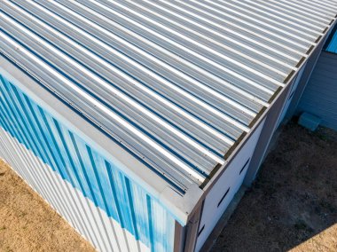 Geniş gri, dik, metal çatılı bir ticari binanın yüksek açılı insansız hava aracı görüntüsü. Perspektif, binanın ölçeğini, bir çatı antenini ve kırsal bir yola ve açık alanlara bitişik konumunu vurguluyor..