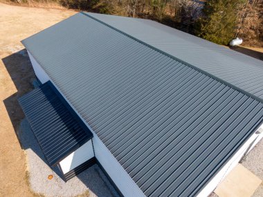 Modern kömür grisi, dik dikişli metal çatının temiz çizgilerini ve pürüzsüz dokusunu gösteren yüksek açılı bir drone görüntüsü. Hava perspektifi, profesyonel kurulum, tepe kapakları ve büyük bir ticari veya hizmet binasının yan hatlarını vurgular.