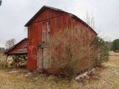 Kuzey Carolina kırsalında, bulutlu bir gökyüzünün altında yıpranmış kırmızı ahşap bir ahır duruyor. Bu kırsal bölgede, harap olmuş bir çiftlik binası ve yakınlarda daha küçük bir kırmızı kulübe var. Amerikan güney mirasının özünü yansıtıyor..