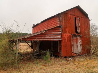 Kuzey Carolina kırsalında, bulutlu bir gökyüzünün altında yıpranmış kırmızı ahşap bir ahır duruyor. Bu kırsal bölgede, harap olmuş bir çiftlik binası ve yakınlarda daha küçük bir kırmızı kulübe var. Amerikan güney mirasının özünü yansıtıyor..