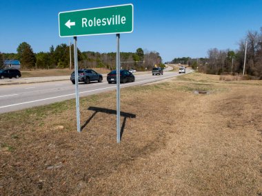 Kuzey Carolina Rolesville 'e doğru giden yeşil bir otoyol işaretinin zemin katındaki görüntüsü. İşaret, çok şeritli bölünmüş bir otoyol boyunca, açık mavi bir gökyüzünün altında, uzaktaki ışık trafiğinin görülebildiği metal direklerde duruyor..