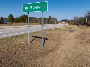 Kuzey Carolina Rolesville 'e doğru giden yeşil bir otoyol işaretinin zemin katındaki görüntüsü. İşaret, çok şeritli bölünmüş bir otoyol boyunca, açık mavi bir gökyüzünün altında, uzaktaki ışık trafiğinin görülebildiği metal direklerde duruyor..