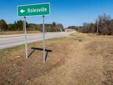 Kuzey Carolina Rolesville 'e doğru giden yeşil bir otoyol işaretinin zemin katındaki görüntüsü. İşaret, çok şeritli bölünmüş bir otoyol boyunca, açık mavi bir gökyüzünün altında, uzaktaki ışık trafiğinin görülebildiği metal direklerde duruyor..