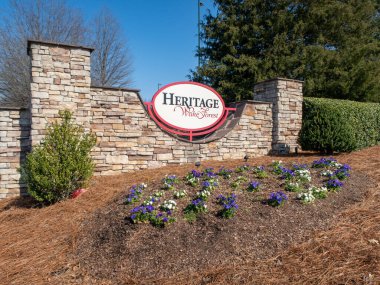 Wake Forest, Kuzey Carolina 'daki Heritage topluluğunun giriş anıtının zemin katlı görüntüsü. Oval tabela, açık mavi gökyüzünün altında mor, beyaz ve yeşil ağaçlarla çevrili istiflenmiş taş bir duvara monte edilmiştir..