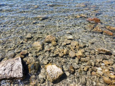 Kristal berraklığında sığ göl suyu yüzeyin altındaki kayaları ve pürüzsüz taşları ortaya çıkararak doğal bir doku oluşturur. Güneş ışığı su boyunca yansıyarak, bu görüntüyü doğa, sağlık, çevre ve arka plan kavramları için ideal hale getirir..