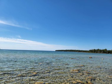 Temiz sığ göl suyu, parlak mavi bir gökyüzü altında sakin bir ufka doğru uzanır. Yüzeyin altında kayalar ve uzak bir ormanlık kıyı şeridi bulunur. Bu barışçıl doğal manzara seyahat, doğa, sağlık ve arka plan kavramları için idealdir..