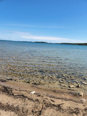 Temiz sığ göl suyu, parlak mavi bir gökyüzünün altında yüzeyin altında görünen taşlarla dolu kumlu bir kıyı şeridiyle karşılaşır. Uzak bir orman ufku bu sakin doğal manzaraya derinlik katar, yolculuk, doğa, sağlık ve geçmiş kullanımı için idealdir..