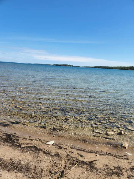 Temiz sığ göl suyu, parlak mavi bir gökyüzünün altında yüzeyin altında görünen taşlarla dolu kumlu bir kıyı şeridiyle karşılaşır. Uzak bir orman ufku bu sakin doğal manzaraya derinlik katar, yolculuk, doğa, sağlık ve geçmiş kullanımı için idealdir..
