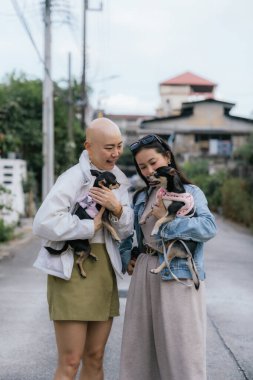 İki neşeli kadın şehir sokaklarında yürürken küçük köpekleri tutuyor.