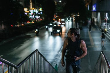 Kameralı ve sırt çantalı bir adam gece şehir merdivenlerinde trafik ışıklarıyla dolaşarak bokeh yaratıyor.