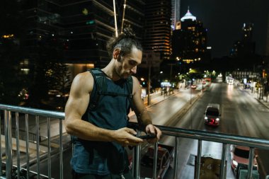 Üstgeçit köprüsünde duran bir adam şehir trafiğine bakarak cep telefonunu kontrol ediyor.