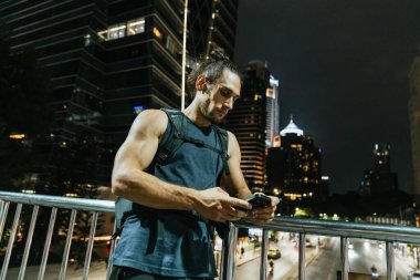 Yaya köprüsünde duran genç adam şehir gecesinde telefonunu kontrol ediyor.