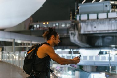 Sırt çantalı bir adam akıllı telefonuyla metro istasyonuna ulaşan treni izliyor.