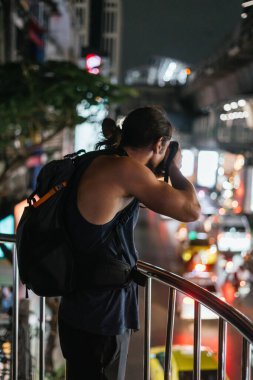 Sırt çantalı bir adam gece şehir ışıklarını ve köprüdeki trafiği yakalamak için kamera kullanıyor.