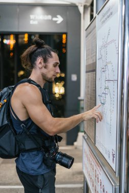 Sırt çantası ve kamerası olan gezgin bir tren istasyonunda tren yolu haritasını inceliyor.