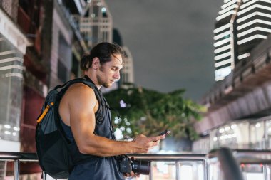 Gezgin gece bir şehir sahnesinde elinde kamerayla telefonunu kontrol ediyor.