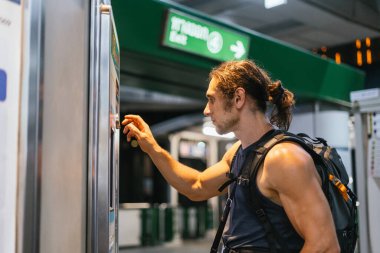 Sırt çantalı bir adam toplu taşıma istasyonunda bilet makinesi kullanıyor.