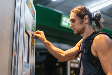 Sırt çantalı bir adam metro istasyonunda bilet otomatı kullanıyor.
