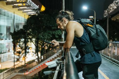 Sırt çantalı bir adam akıllı telefonuyla şehrin sokaklarında gezinirken tırabzanlara yaslanıyor.