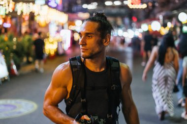 Kameralı ve sırt çantalı bir adam renkli bir gece marketinde yürüyor.