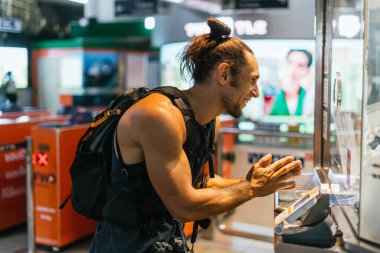 Sırt çantalı bir adam toplu taşıma istasyonunda otomatik bilet gişesi kullanırken gülümsüyor.