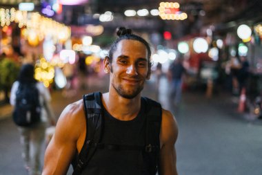 Gece aydınlık bir sokakta sırt çantasıyla gülümseyen erkek turist.