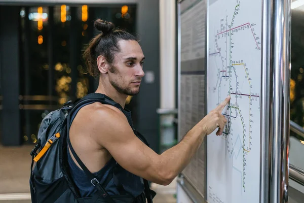 Sırt çantalı bir adam metro haritasına bakıyor, tren istasyonunda yolculuğunu planlıyor.
