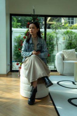 Genç bir kadın bir puf üzerinde oturuyor, çevreyi gözlemliyor, modern bir evde akıllı telefon tutuyor.