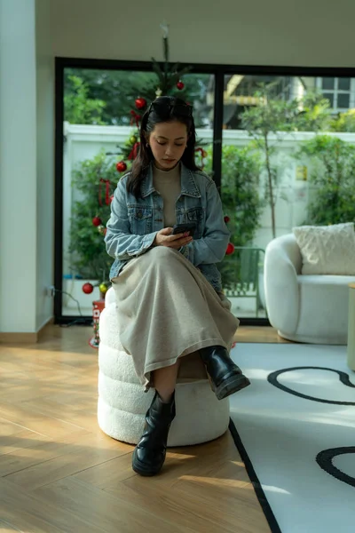 Taburede oturan genç bir kadın modern bir oturma odasında cep telefonunu kontrol ediyor.