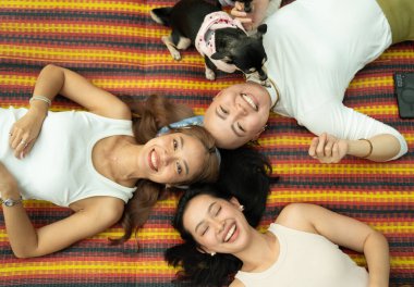 Üç gülümseyen kadın ve köpekleri renkli bir hasırda dinleniyorlar.