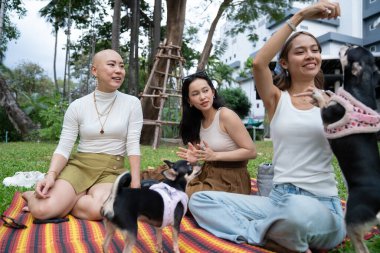 Kadın arkadaşlar yeşil bir parkta açık hava pikniğinde köpekleriyle dinleniyorlar.