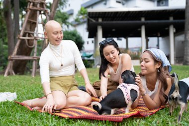 Üç arkadaş parkta evcil köpekleriyle çimlerin üzerinde piknik yapıyorlar.