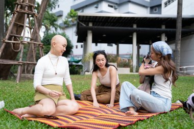 Üç kadın ve bir köpek parkta çimlerin üzerinde pikniğin tadını çıkarıyorlar. Arkadaşlık ve destek sağlıyorlar.
