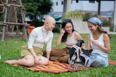 Üç gülümseyen kadın ve parkta bir piknik minderinde dinlenen iki küçük köpek.