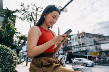 Genç bir kadın gülümsüyor, hareketli bir şehir sokağında mobil teknolojiden zevk alıyor.