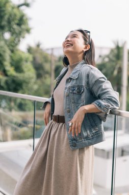 Genç bir kadın gülümsüyor, açık hava balkonunda kot ceket giyiyor.