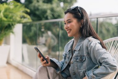 Gülümseyen genç bir kadın balkonda rahatlıyor, cep telefonuyla bağlantı kuruyor.