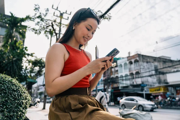 Genç bir kadın gülümsüyor, hareketli bir şehir sokağında mobil teknolojiden zevk alıyor.