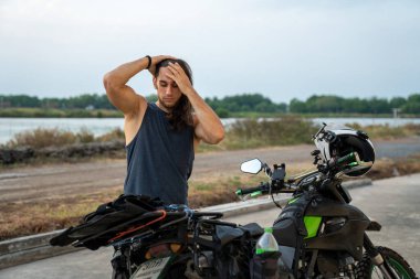 Uzun saçlı bir adam maceralı bir yolculukta motosikletinin yanında dinleniyor.