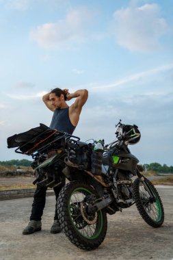 Uzun saçlı bir adam, seyahat için yüklü çift spor bir motosikletin yanında geriniyor.