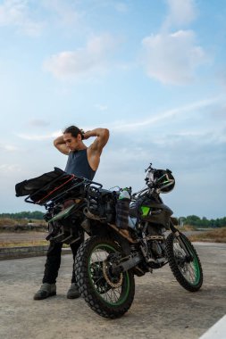 Macera motosikletinin yanında duran, esneyen ve yolculuğa hazırlanan bir adam.