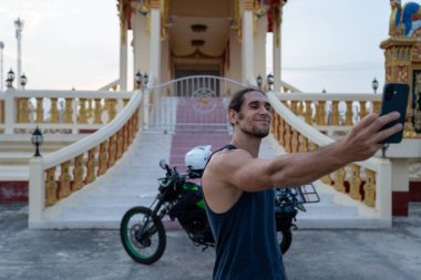Adam gülümsüyor, cep telefonu tutuyor ve Asya 'nın güneydoğusundaki bir tapınakta selfie çekiyor.