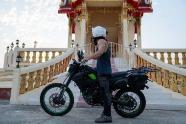 Kasklı bir adam Tayland tapınağının önünde motosiklet macerasının yanında duruyor.