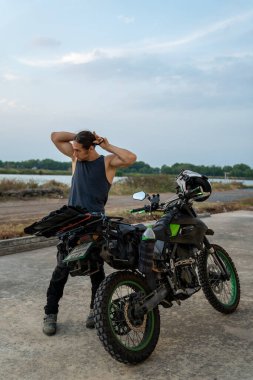 Uzun saçlarını arkadan bağlayan bir adam, açık havada dolu bir spor motosikletinin yanında duruyor.