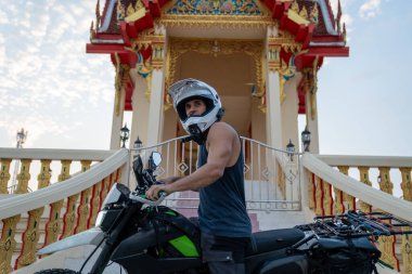 Motosikletli bir adam süslü bir Budist tapınağının önünde kask takıyor.