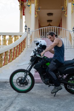 Sıradan giyimli bir adam kasklı bir motosiklete biniyor. Geleneksel Tayland mimarisinin yanına park etmiş.