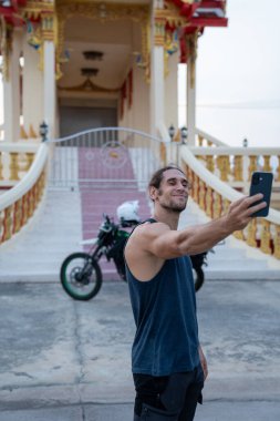 Gülümseyen adam Tayland tapınağının önünde motosikletiyle selfie çekiyor.