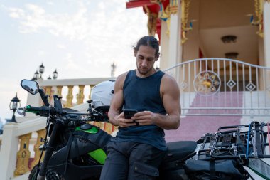 Adam renkli bir Tayland tapınağının önünde motosikletle seyahat ederken akıllı telefonu kontrol ediyor.
