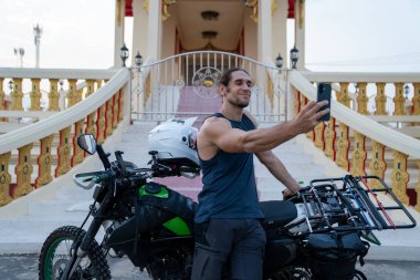 Gülümseyen bir adam, Tayland tapınağının önünde macera motosikletiyle selfie çekiyor.